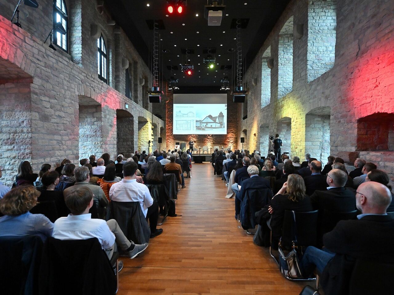 Blick in den voll besetzten Saal mit großer Leinwand und Projektion